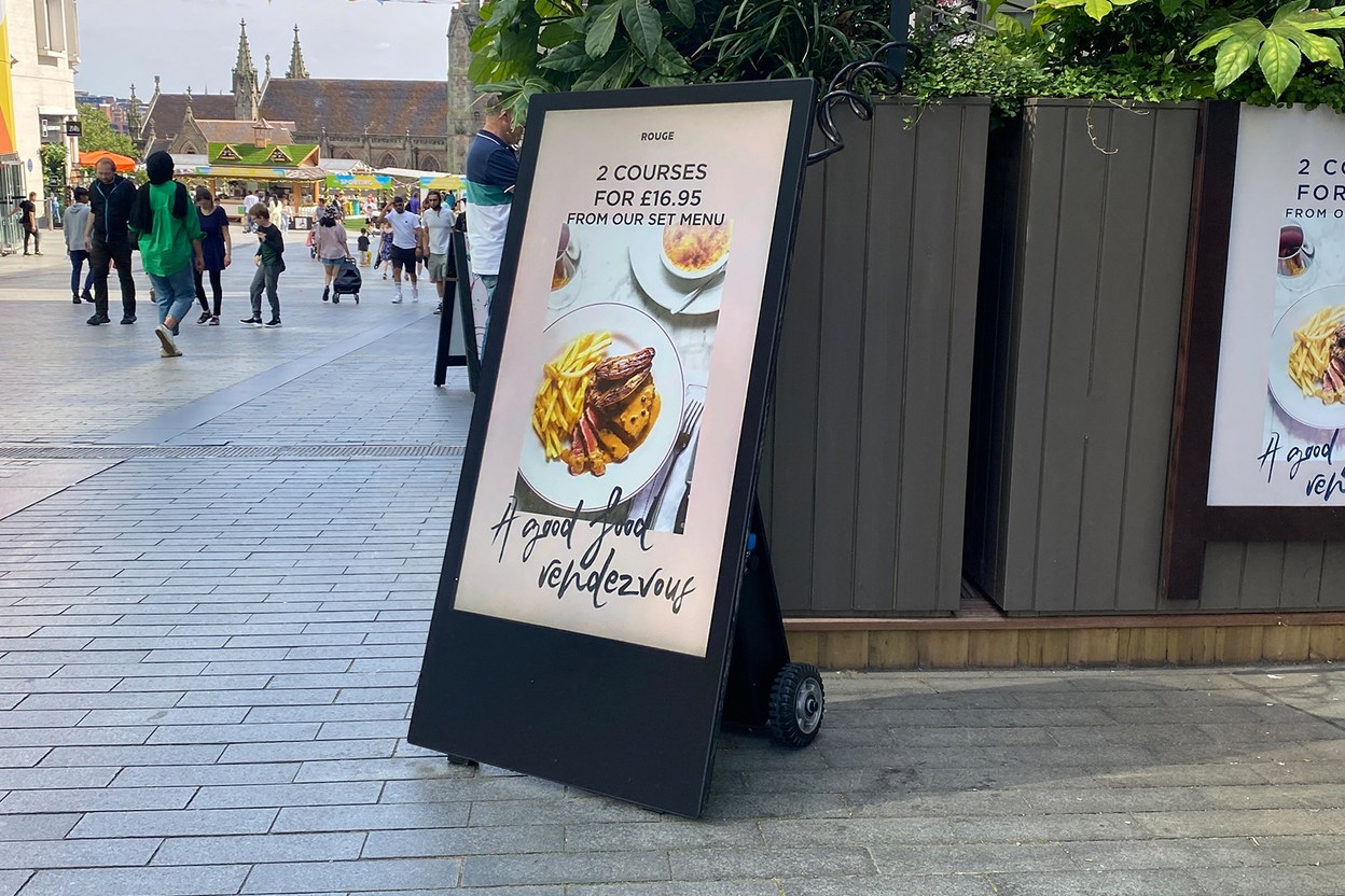 Chevalet numérique sur roulettes posé sur le trottoir d'une rue piétonne, écran vertical diffusant le menu d'un restaurant